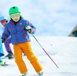 Picture of a child wearing a blue plaid coat, yellow snow pants, a green hat, and goggles. They are wearing skis and are learning to ski.
