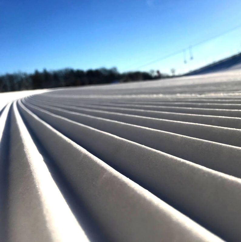 Picture of the freshly groomed snow on the hill at Pine Knob Resort. The sky is blue and the snow is sparkling and white. There are trees off in the background.