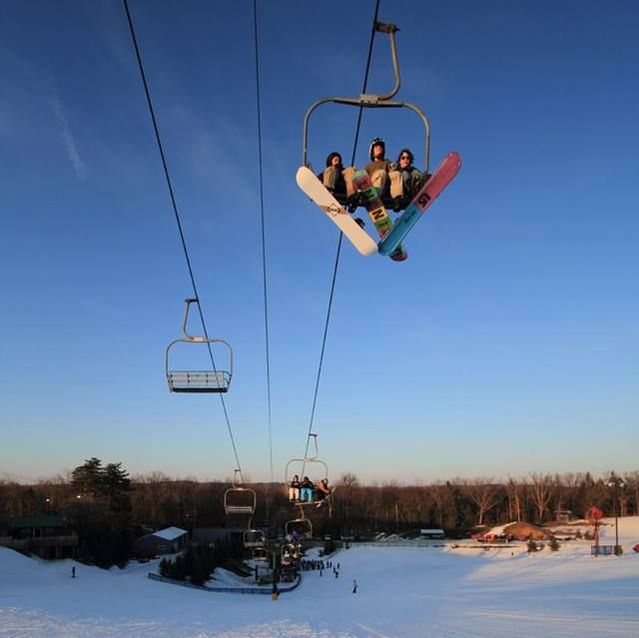 Picture of guests riding a chair lift up the hill at Pine Knob. The picture was taken from a higher vantage point on the hill so you can see the chairlift, blue sky, snow on the hill, and buildings and trees at the bottom of the hill.